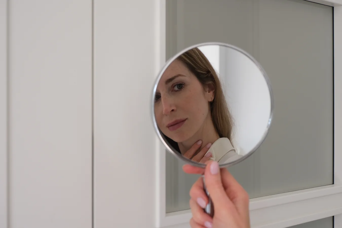 A woman looks at her face in a round hand mirror in front of a white cabinet.