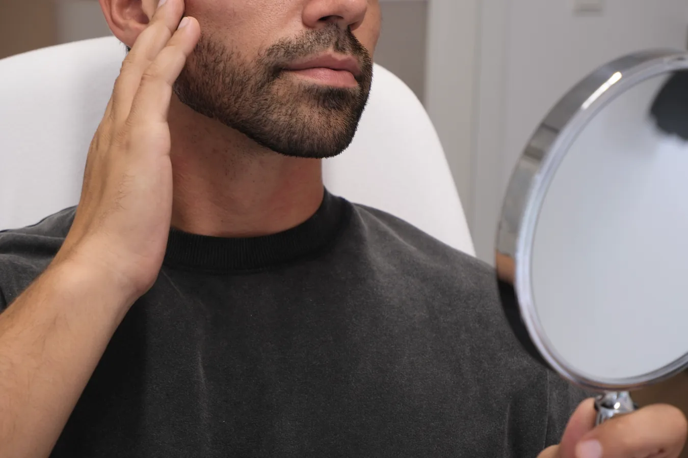 Bearded man looking at his face in a hand mirror after a treatment and touching his cheek.