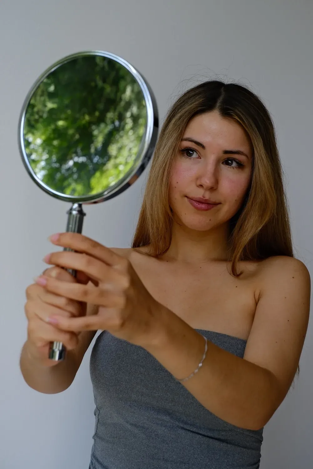 Woman with long hair holding a round hand mirror and looking at the result of her treatment at Skinthetics