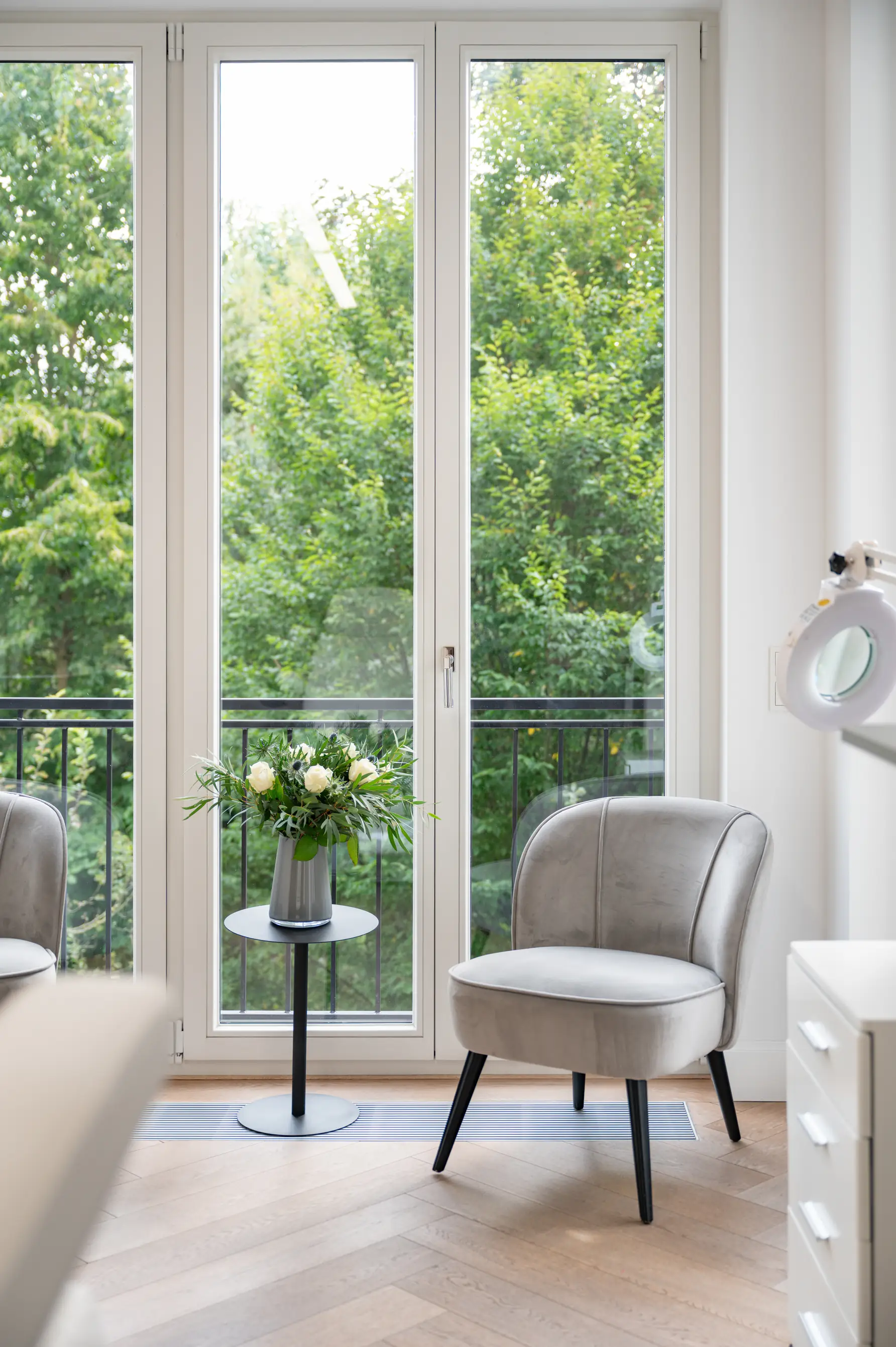 Consultation room interior:
Modern room with a gray armchair, a small table with flowers and large windows overlooking green trees.