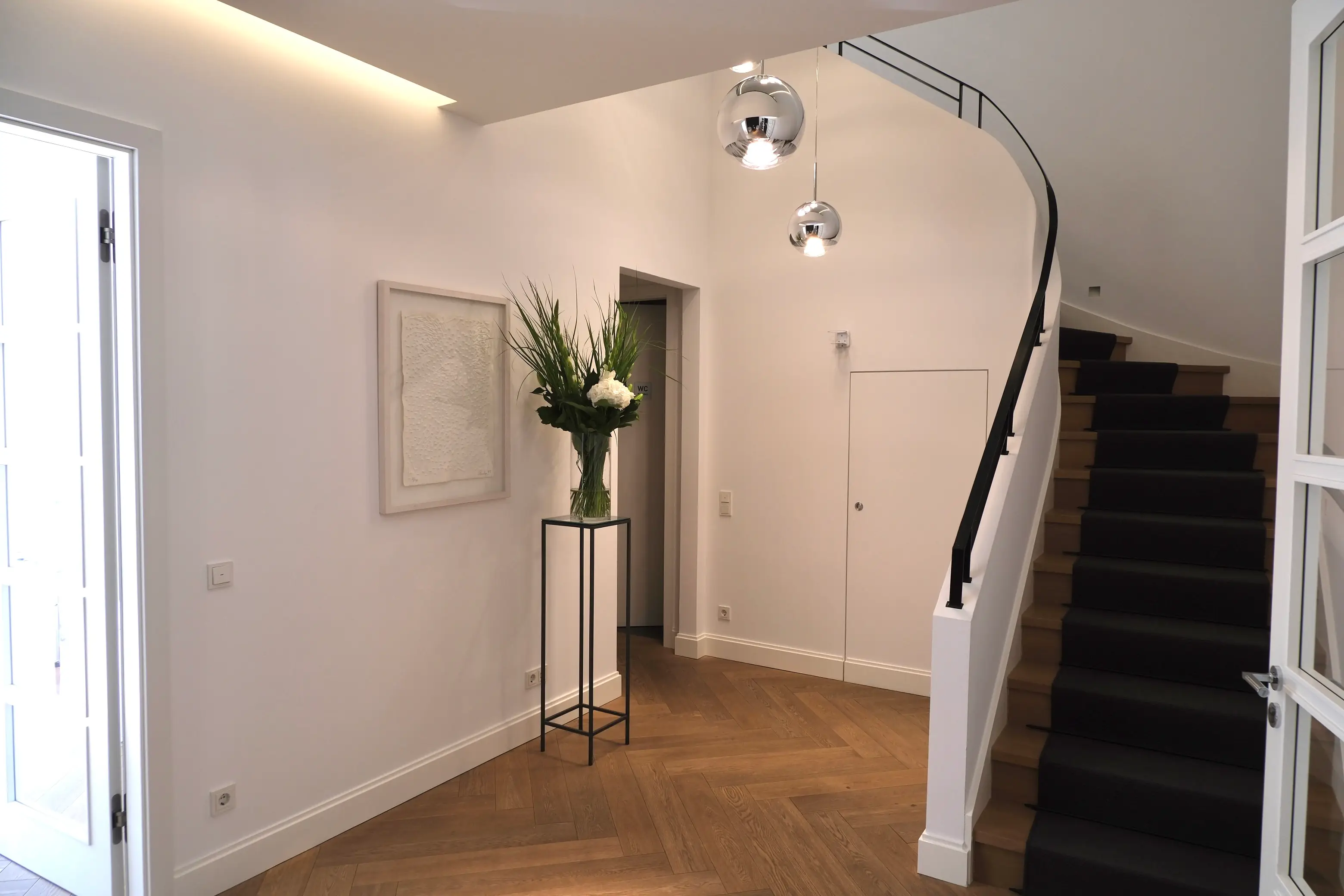 Cosmetic institute interior:
Modern, bright hallway with wooden parquet flooring, a staircase with a black carpet runner, a large flower arrangement on a narrow black stand and two hanging globe lights.