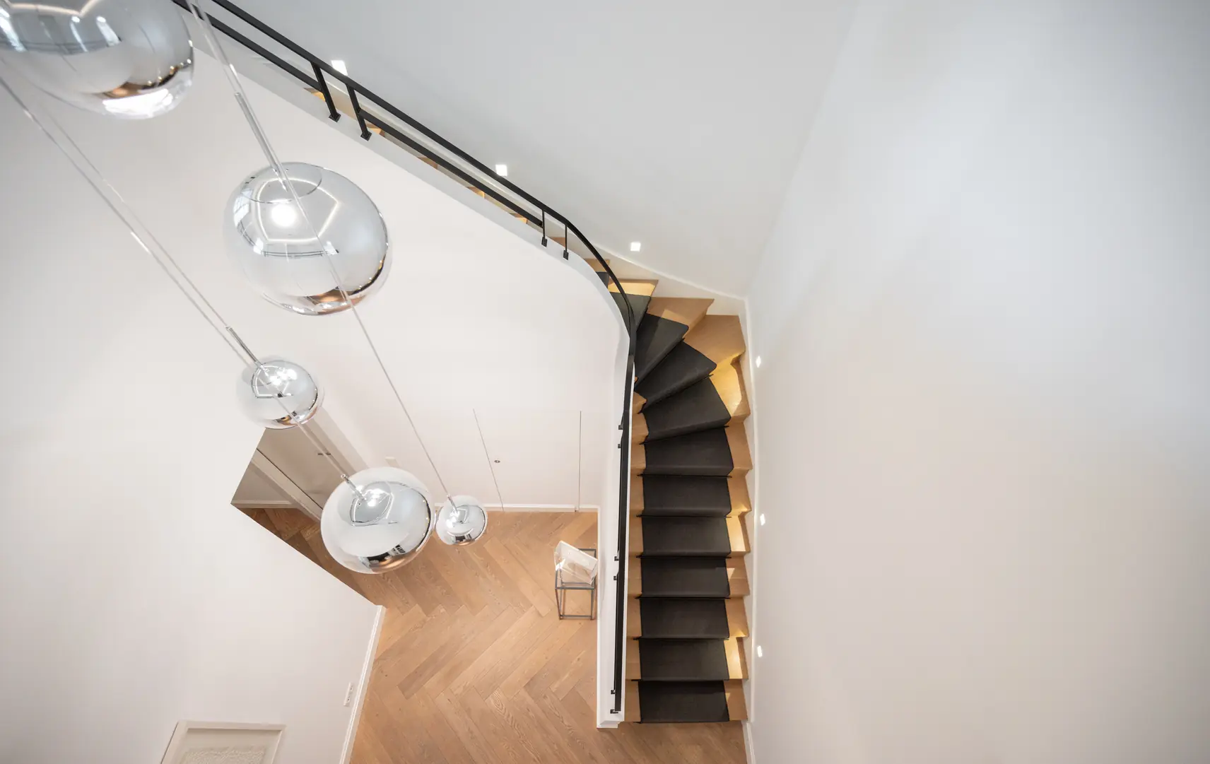 Modern wooden staircase with black carpet and hanging silver globe lights in a white room.