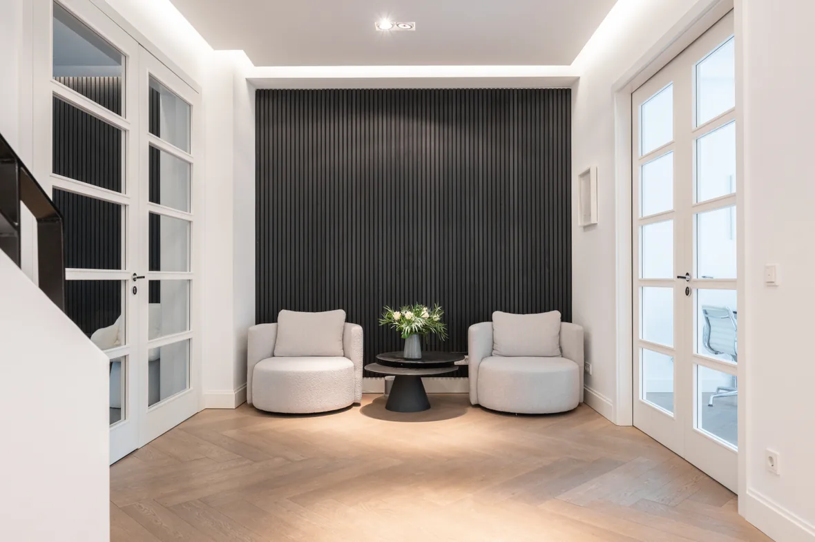 Modern waiting area with two beige armchairs, a black table and a vertical black wall panel.