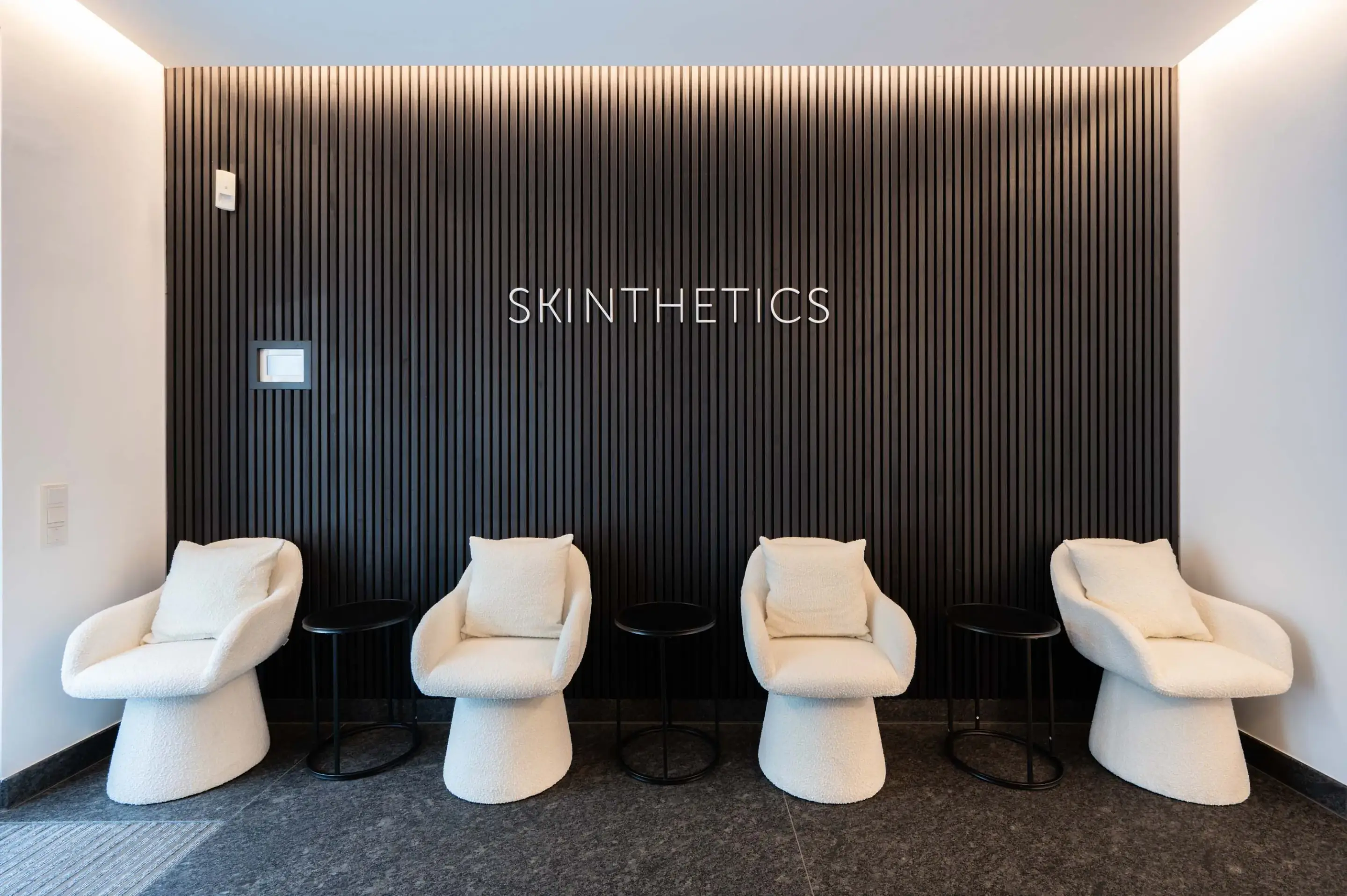 Waiting room: four cozy white chairs with cushions in front of a black, vertically fluted wall with the lettering "SKINTHETICS".